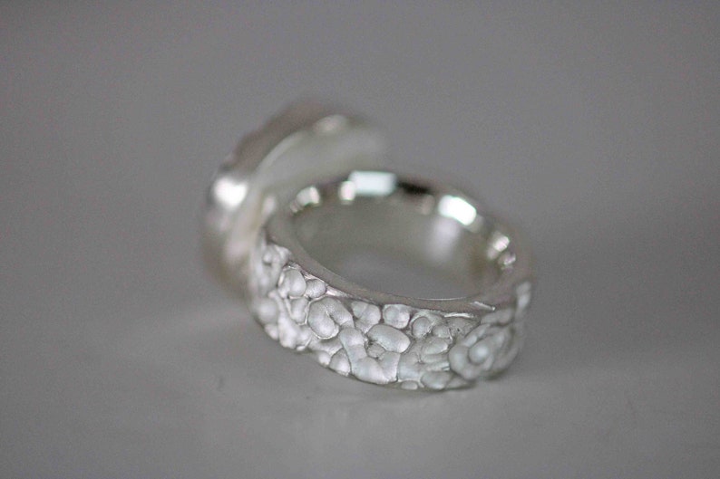 May include: Close-up of a silver ring with a textured band and a smooth, rounded top. The ring's band features a detailed, organic pattern, and the overall design has a handcrafted appearance. The ring is likely made of sterling silver.