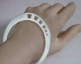 May include: A silver bangle bracelet with a geometric design. The bracelet is open and has a smooth, brushed finish.