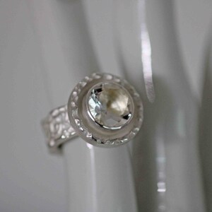 May include: A silver ring with a large, clear gemstone at the center. The gemstone is surrounded by a decorative, circular setting. The band of the ring has a textured, floral design.