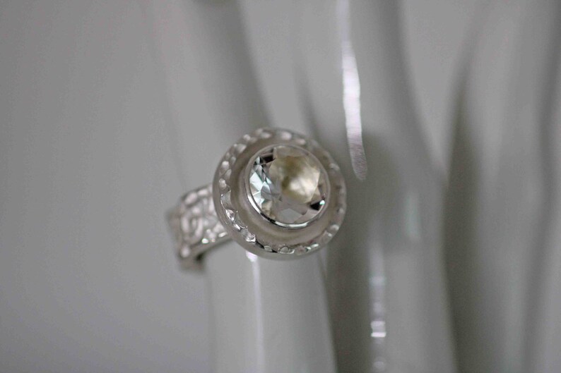 May include: A silver ring with a large, clear gemstone at the center. The gemstone is surrounded by a decorative, circular setting. The band of the ring has a textured, floral design.