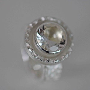 May include: A close-up of a silver ring with a large, clear, faceted gemstone. The gemstone is round and surrounded by a halo of smaller, clear stones. The ring has a detailed, textured band.