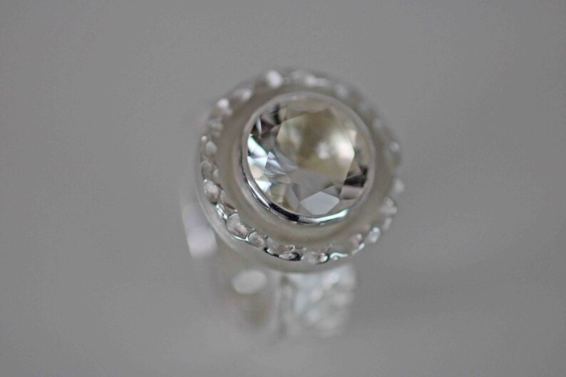 May include: A close-up of a silver ring with a large, clear, faceted gemstone. The gemstone is round and surrounded by a halo of smaller, clear stones. The ring has a detailed, textured band.