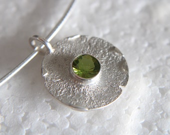 Op de afbeelding: Een zilveren hanger met een getextureerd oppervlak en een groene edelsteen in het midden.