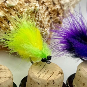 May include: Five different fly fishing lures with black, red, orange, yellow, and purple feathers. The lures are mounted on cork.