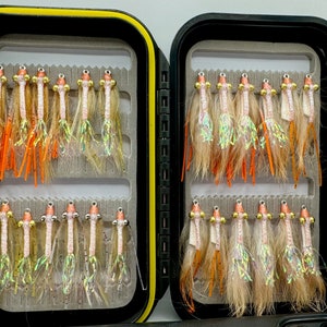 May include: A black fly fishing box with two rows of fly fishing lures. The lures are tied with orange, white, and brown feathers. The box is open and the lures are visible.