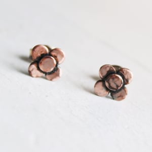 May include: A pair of copper flower-shaped stud earrings. The earrings have a hammered texture and a warm, rose gold tone.