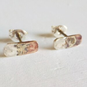 May include: A pair of silver stud earrings with a rectangular shape. The earrings have a textured surface with a mix of silver and copper tones.