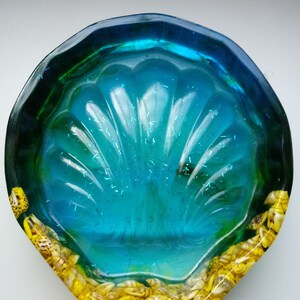 May include: A scalloped shell-shaped bowl made of translucent blue and green resin with embedded silver glitter. The bottom edge of the bowl is decorated with a dense arrangement of small, yellow and brown patterned seashells.