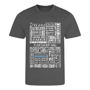 May include: A charcoal grey t-shirt with a white text design. The design is a vertical rectangle of song titles from the band Fleetwood Mac, including "Dreams," "Rhiannon," and "Go Your Own Way."