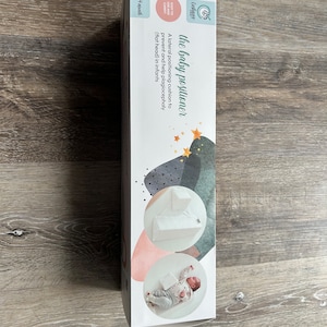 May include: White baby positioning cushion with a star pattern on the box. The text on the box reads "the baby positioner" and "A lateral positioning cushion to prevent and help plagiocephaly (flat head) in infants".