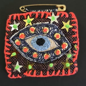 May include: A square fabric patch featuring an embroidered blue eye motif at the center, surrounded by gold star-shaped sequins and small red circular accents. The patch has a bright orange stitched border and a gold safety pin attached at the top.