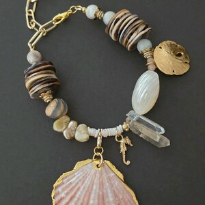 May include: A handcrafted necklace featuring a pink seashell pendant with gold trim, complemented by a seahorse charm, quartz crystals, and various beads in neutral tones. The necklace has a gold chain and clasp.