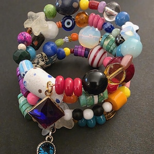 May include: A multi-strand bracelet with a variety of colorful beads. The bracelet features a mix of shapes, including round, star, and geometric beads in shades of blue, pink, green, and yellow. A blue gem pendant hangs from the bracelet.