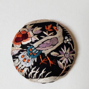 May include: A round, black button with a white, purple, orange, and teal embroidered design of a bird and flowers.