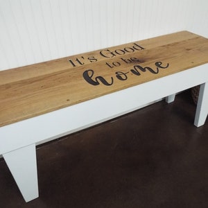 May include: White wooden bench with a natural wood top. The top has the words "It's Good to be home" carved into it.