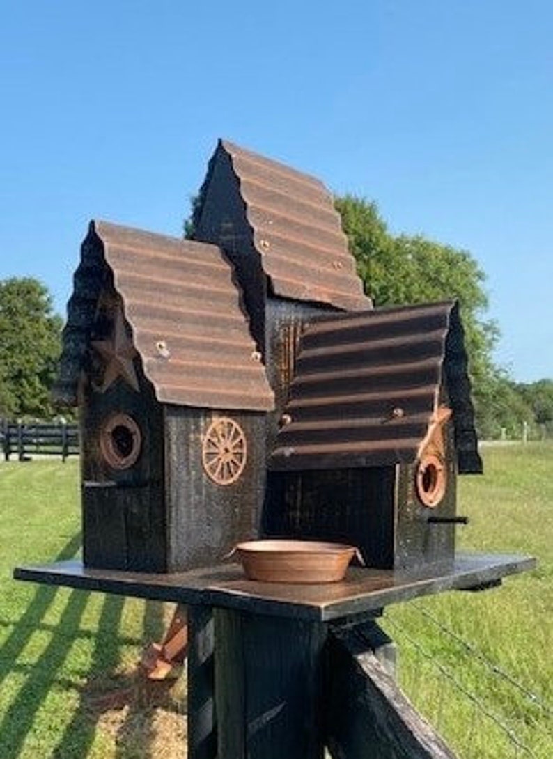 May include: Three rustic birdhouses made of metal and wood, painted black with brown accents. The birdhouses are stacked on a wooden post and have a small metal dish in front of them.