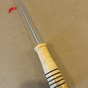 May include: A metal latch hook tool with a long, thin silver shaft and a curved tip. The handle is made of light-colored wood with a smooth, polished finish and features four distinct horizontal black rings etched into the grip area.