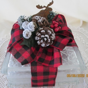 May include: A decorative glass block adorned with a red and black buffalo plaid bow, pinecones, and frosted berries. A metal moose silhouette sits atop the arrangement, creating a rustic, festive accent. The block is perfect for seasonal decor.