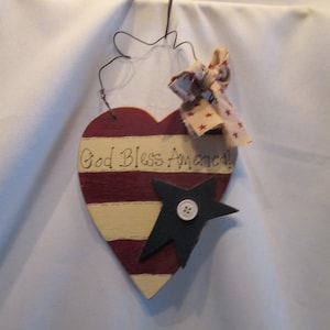 May include: A wooden heart-shaped sign with red and white stripes and the text "God Bless America!" painted on it. A black star with a white button in the center is attached to the sign. A red, white, and blue ribbon is tied to the top of the sign.