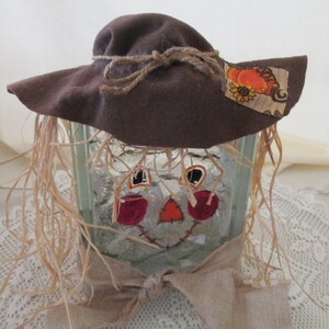 May include: A scarecrow-themed glass block with a brown felt hat, straw hair, and a burlap bow. The face is painted with red cheeks, a black stitched mouth, and orange nose.