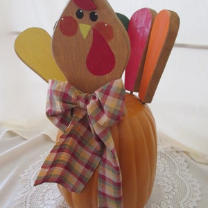 May include: A wooden turkey figurine with a pumpkin body, red, orange, and yellow feathers, and a plaid bow. The turkey has a painted face with a red beak and black eyes.