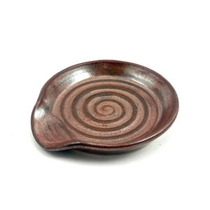 以下が含まれることがあります： 茶色と赤色の手作りのセラミック製スプーンレスト。 スプーンレストは中央に渦巻き模様があり、食器を置くための湾曲したエッジがあります。 キッチン用品は白い背景にあります。