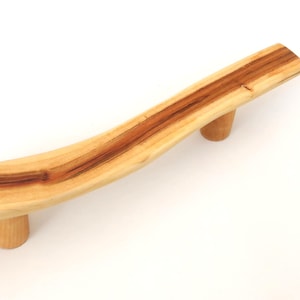 May include: A handcrafted wooden serving tray with a natural, curved design. The tray is made of light-colored wood with a prominent, dark brown grain running through the center. Two cylindrical wooden feet provide support.