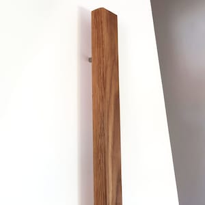 May include: A vertical, rectangular wooden door pull with a natural wood grain finish. The pull is mounted on a white surface with two small, silver-colored screws. The wood is a warm brown color.