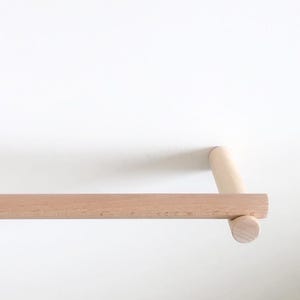 Könnte beinhalten: Eine Handtuchstange aus hellem Holz mit einer langen, horizontalen zylindrischen Stange, die an beiden Enden von zwei kleineren zylindrischen Wandhalterungen getragen wird und eine glatte Oberfläche mit natürlicher Holzmaserung aufweist.