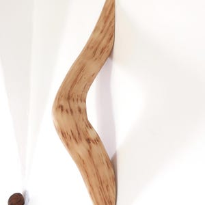 Peut inclure: Un boomerang en bois brun clair de forme incurvée, appuyé contre un mur blanc. Le grain du bois est visible, avec des stries brun foncé. Un petit objet rond et brun est posé sur le sol près du mur.