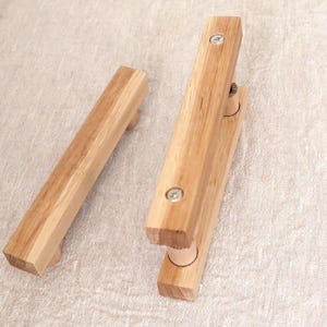 May include: Two light-colored wooden cabinet handles with a rectangular profile. One handle is positioned horizontally, while the other is shown with two silver-toned metal screws visible on the top surface, securing the handle to its mounting posts.