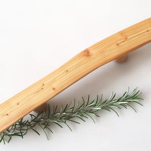 Könnte beinhalten: Ein hellfarbener Holzgriff mit geschwungenem Design, möglicherweise für einen Schrank oder eine Schublade. Der Griff ist diagonal positioniert, darunter ein Rosmarinzweig. Die Holzmaserung ist sichtbar, und der Griff hat ein natürliches, rustikales Aussehen.