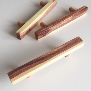 Op de afbeelding: Drie houten kastgrepen. Elke greep is rechthoekig met een lichte houtkleur aan één kant en een donkerdere, roodbruine kleur aan de andere kant. De grepen hebben kleine, ronde, lichtgekleurde houten knoppen aan elk uiteinde.
