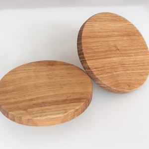 May include: Two round wooden coasters with a natural wood grain pattern. The coasters are light brown and have a smooth, finished surface. One coaster is lying flat, while the other is tilted slightly.
