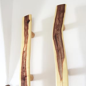 Könnte beinhalten: Zwei Türgriffe aus Holz mit einer hellen Außenseite und einem dunkelbraunen Maserungsmuster. Sie sind an einer weißen Wand mit kleinen, runden Holzstiften befestigt. Hergestellt aus Naturholz.