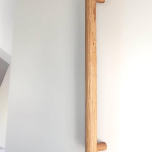 May include: A long, wooden cabinet door pull with a cylindrical shape and rounded edges. The pull is made of natural wood with a light brown color and a visible wood grain.