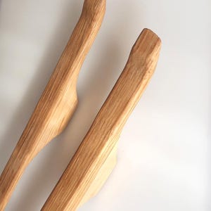 Könnte beinhalten: Zwei Salatgabeln aus Holz mit natürlicher Maserung. Die Zangen haben gebogene Griffe und breite, flache Enden. Das Holz ist hellbraun und die Zangen scheinen handgefertigt zu sein.