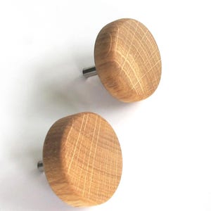 May include: Two wooden cabinet knobs with a natural oak finish. Each knob is round with a flat face and a silver-colored metal stem. The knobs are set against a white background.