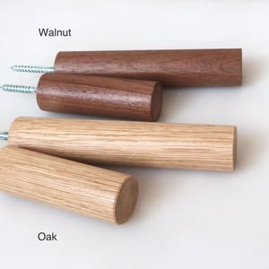 Op de afbeelding: Twee cilindrische houten wandhaken met het label Walnut, met een donkerbruine nerf, en twee cilindrische houten wandhaken met het label Oak, met een lichtbruine nerf. Elke haak is voorzien van een metalen schroef aan de achterkant.