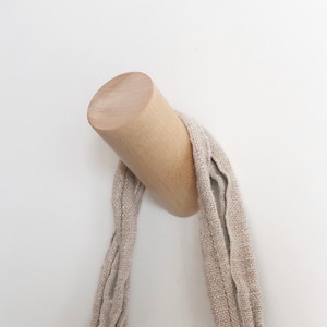 Könnte beinhalten: Ein helles Wandhaken aus Holz mit zylindrischem Design. Eine beige Stoffschlaufe hängt an dem Haken und erzeugt eine schlichte, minimalistische Ästhetik. Der Haken ist an einer weißen Wand befestigt.