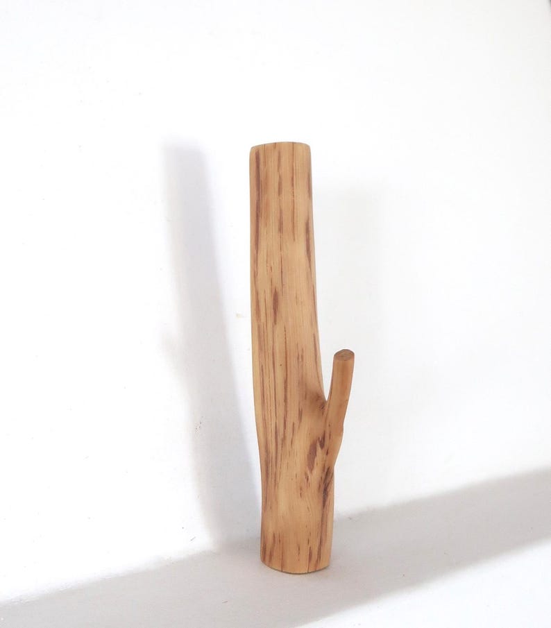Puede incluir: Un gancho de pared de madera vertical con forma de segmento de rama de &aacute;rbol. La madera de color marr&oacute;n claro presenta vetas naturales de color marr&oacute;n oscuro y un solo pomo que sobresale en el lateral para colgar objetos. La superficie es lisa y cil&iacute;ndrica.