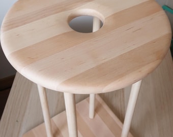 May include: A light-colored wooden stand featuring a circular top with a central hole, supported by four slender cylindrical legs attached to a square base. The entire structure is constructed from joined wooden planks with a smooth finish.