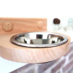 Puede incluir: Un cuenco de madera para mascotas con un inserto de acero inoxidable. El cuenco está montado en un estante de madera de color claro con dos clavijas de madera. El diseño es simple y moderno, adecuado para gatos o perros pequeños.