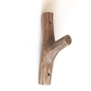 Puede incluir: Un gancho de pared fabricado a partir de una sola pieza de madera marr&oacute;n natural con una textura de grano visible. El gancho presenta un cuerpo principal vertical con dos orificios de montaje circulares y una rama inclinada.