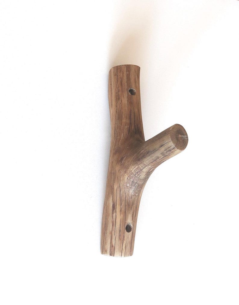 Puede incluir: Un gancho de pared fabricado a partir de una sola pieza de madera marr&oacute;n natural con una textura de grano visible. El gancho presenta un cuerpo principal vertical con dos orificios de montaje circulares y una rama inclinada.