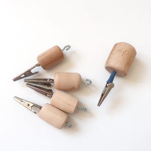 May include: Five craft clips featuring cylindrical light-colored wooden handles. Each clip has a metal alligator-style jaw at one end and a small metal hook at the opposite end. One clip includes a blue sleeve connecting the metal jaw to the wood.
