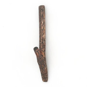 Puede incluir: Un gancho de pared de madera marr&oacute;n con forma de peque&ntilde;a rama de &aacute;rbol y acabado de corteza texturizada. El gancho presenta un tallo principal vertical y una rama m&aacute;s corta e inclinada en el lateral para colgar diversos objetos.