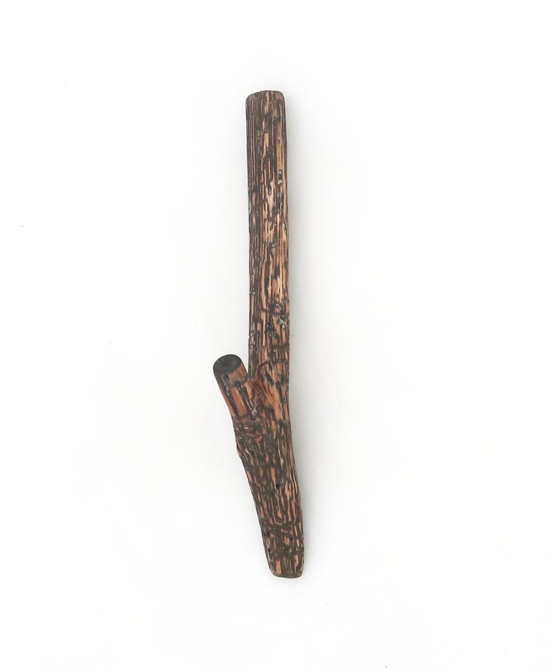 Puede incluir: Un gancho de pared de madera marr&oacute;n con forma de peque&ntilde;a rama de &aacute;rbol y acabado de corteza texturizada. El gancho presenta un tallo principal vertical y una rama m&aacute;s corta e inclinada en el lateral para colgar diversos objetos.