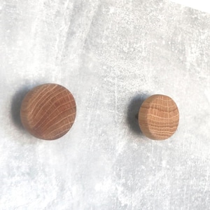 May include: Two round wooden knobs with a natural wood grain pattern. The knobs are light brown and appear to be made of oak. They are mounted on a light gray surface.