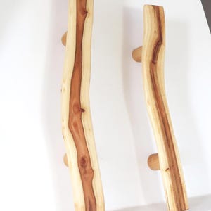 May include: Two wooden door pulls with a natural, organic design. The pulls are made from light-coloured wood with a darker, central grain. Each pull has a small, rounded knob for gripping. The numbers 1 and 2 are printed at the bottom.